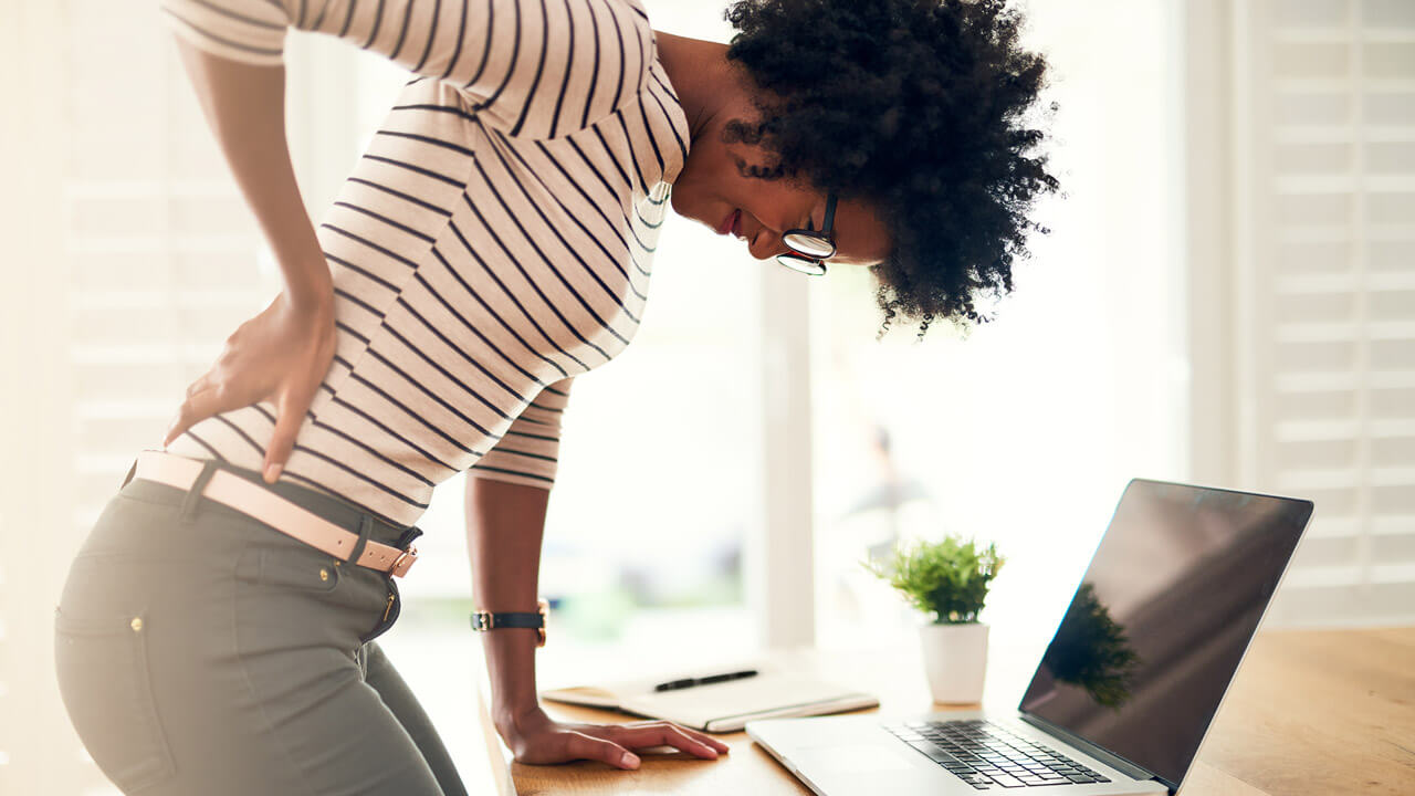 A Woman Holds Her Back While Leaning On A Desk In Front Of A Laptop