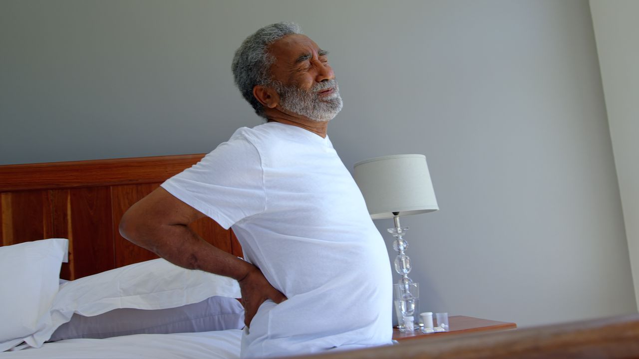 A Man In A White Shirt Is Sitting On A Bed Holding His Back In Pain
