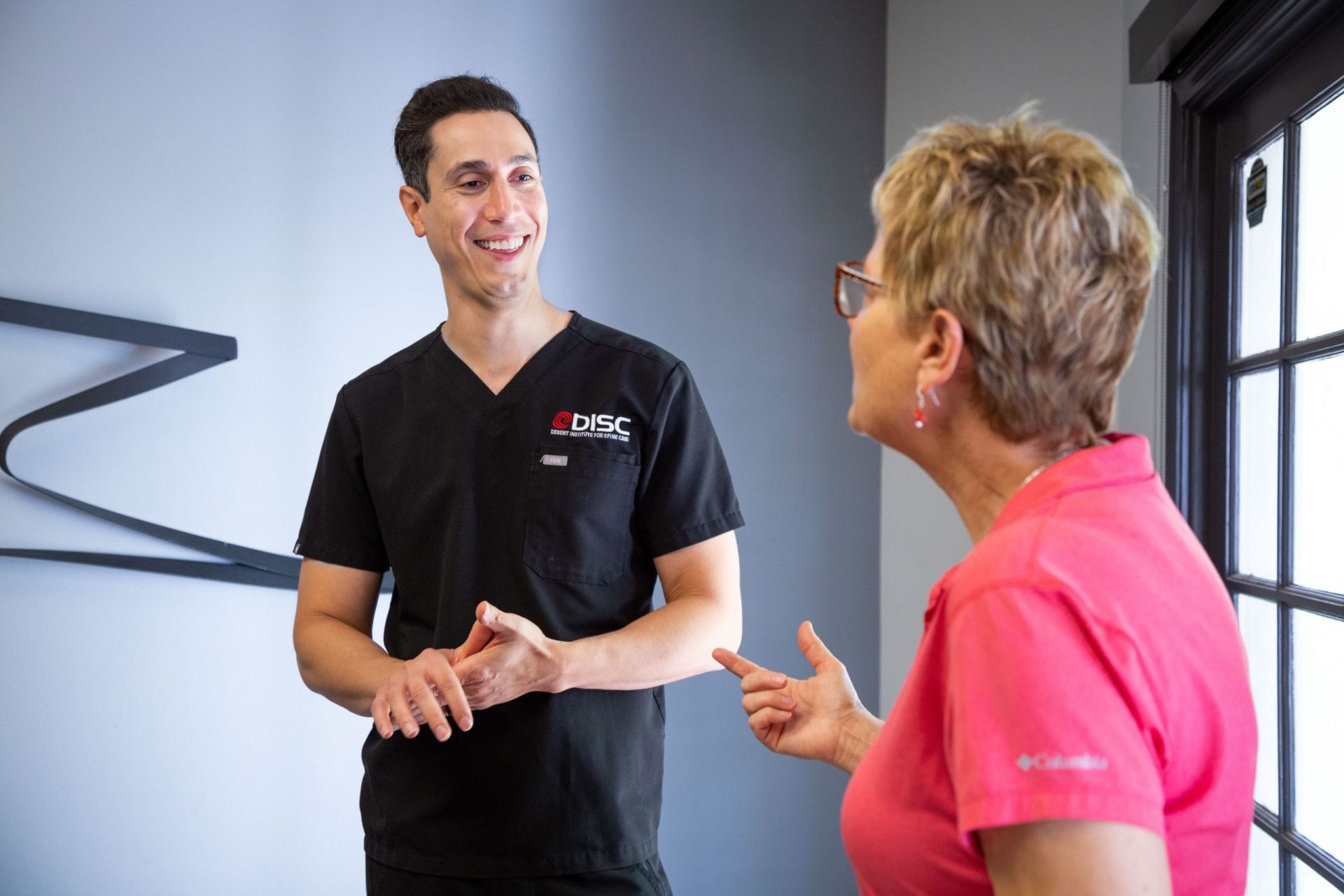 Dr. Salari Wearing A Black Disc Scrub Talking To A Woman In A Pink Shirt